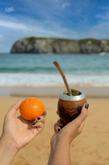 Woman's hands holding a tangerine and a yerba mate drink on the beach