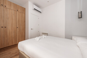 Fototapeta na wymiar Bedroom with double bed with white bedding, wardrobe with chests and oak wood doors and white air conditioner on the wall