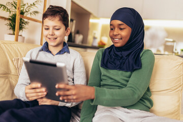 Muslim girl and her caucasina friend sitting on sofa at home using digital tablet.