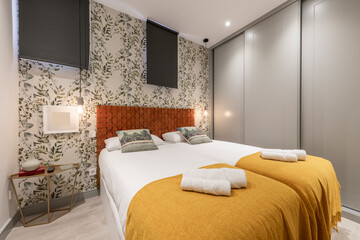 Bedroom with double beds with wall covered in decorative wallpaper, intertwined brown rope headboard, wooden gray sliding door wardrobe and mustard colored blankets