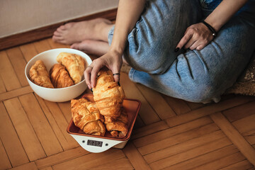 a young girl sits on the floor and tries to control her weight and keep track of calories, she weighs croissants on a baking scale. The concept of the problem of excess weight.