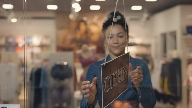 Young african american woman turning a sign from closed to open on the door of a women's brand clothing store in a mall. The clothing store starts its working day. Close up. Slow motion.