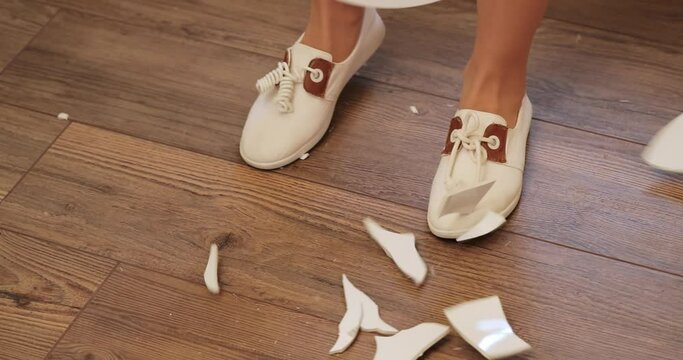 A white plate falls and breaks into small pieces in the kitchen against the background of female legs in shoes. the girl breaks the plate on the floor. view from above
