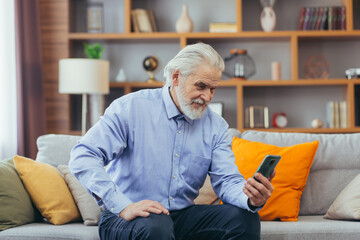Senior man at home sitting on sofa looking at phone screen, talking on video call with family