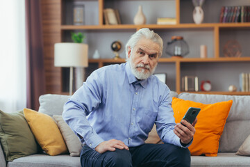 Senior man at home sitting on sofa looking at phone screen, talking on video call with family