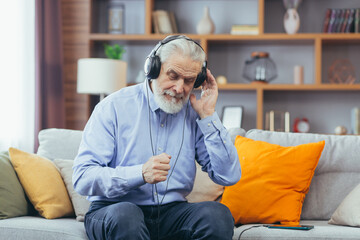 Obraz premium Senior gray-haired man sitting on sofa at home, relaxing and listening to music on headphones, using music app on phone