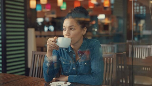 View through a glass showcase of a young African American woman enjoying coffee in a mall cafe. A woman is resting after shopping and waving to her friend passing by. Close. Slow motion.