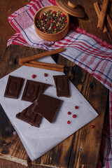 Broken bar of dark bitter chocolate with chilly pepper, red pea pepper, cinnamon flavour on paper, wooden board background. Unusual sweets taste