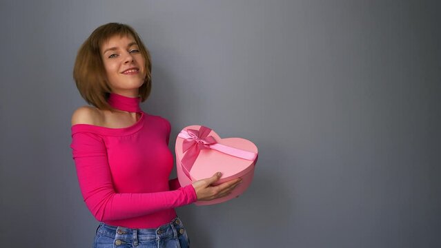 Attractive caucasian woman in pink bodysuit showing heart shaped gift bpx in her hands in front of gray wall, copy space.