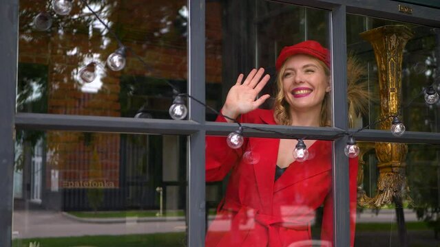Stylish cheerful blonde woman in red suit and red capis waving her hand to somebody through the cafe veranda window.