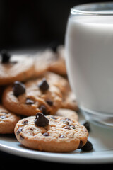Chocolate chip cookies on a white plate, with a cold glass of milk on the side, over a wooden table. Side view macro photography vertical image