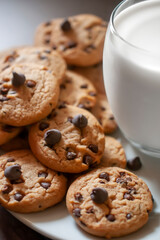 Chocolate chip cookies on a white plate, with a cold glass of milk on the side, over a wooden table. Vertical image