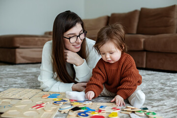 Mother and little daughter play educational games on rug in room. Toddler baby in a brown sweater.