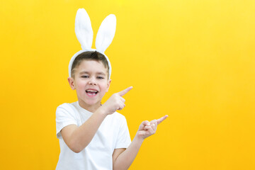 cheerful boy with rabbit ears on his head on a yellow background. Funny happy child points his finger at an empty space. copy space for text, layout