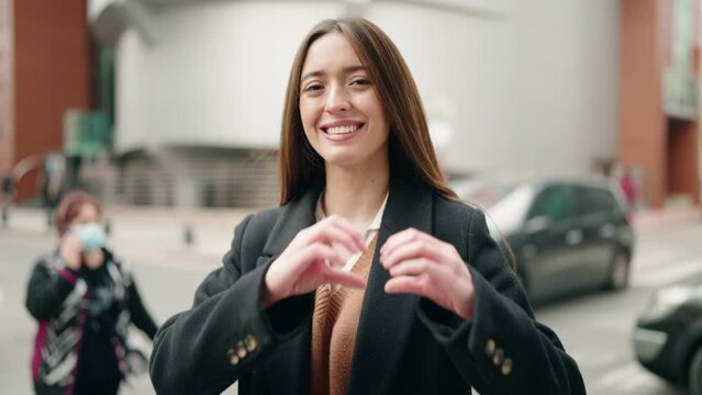 Young hispanic woman smiling confident doing heart gesture with hands at street