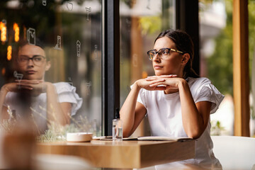 A thoughtful woman is sitting next to the window in the cafeteria and looking through it. 