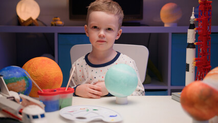 Portrait smiling little blond boy sitting at home at table near planet Uranus around many planets...