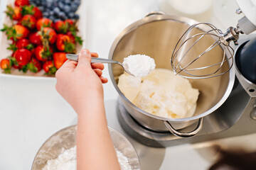 top view. A cook adds a spoonful of flour to prepare dough in a mixer bowl.
