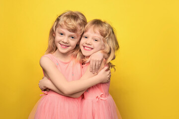 Portrait of twin girls. Two cute attractive cheerful girls of preschool age. Smiling sisters hugging and looking at the camera. Curly blonde 5 years old in a pink dress on a bright yellow background.