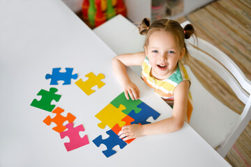 A laughing sweet girl is sitting at a table and assembling a puzzle of colorful elements on autism Day.  Logical thinking and development. The child looks up into the camera. 