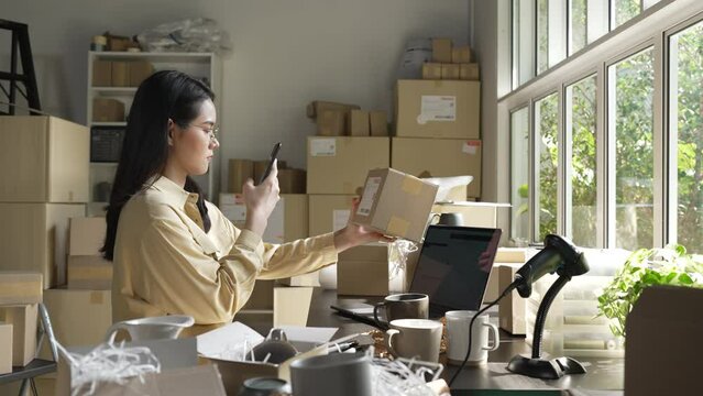 Asian female small business owner using mobile app on smartphone checking parcel box. Warehouse worker, seller holding phone scanning retail dropshipping package postal parcel bar code.
