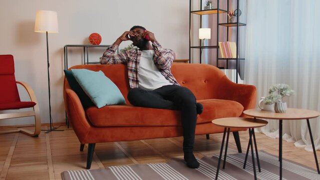 Portrait of tired sleepy man in shirt talking on mobile phone with friend at home. Sad African American guy freelancer making online conversation speaks on smartphone sitting on sofa at living room