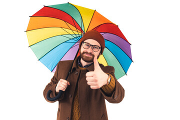 Obraz na płótnie Canvas Portrait of young attractive Caucasian man holding a multicoloured umbrella and showing thumb up to the camera positivity optimism concept brown casual attire isolated white background studio shot