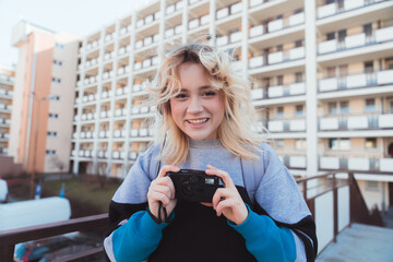 Grinning caucasian girl holding black old-fashioned camera. Block of flats scenery. 90s vibes. High quality photo