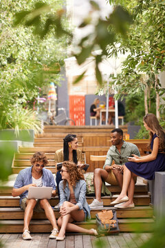 Chilling on campus between classes. A group of young students relaxing outdoors with technological devices.