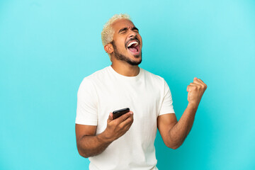 Young Colombian handsome man isolated on blue background with phone in victory position