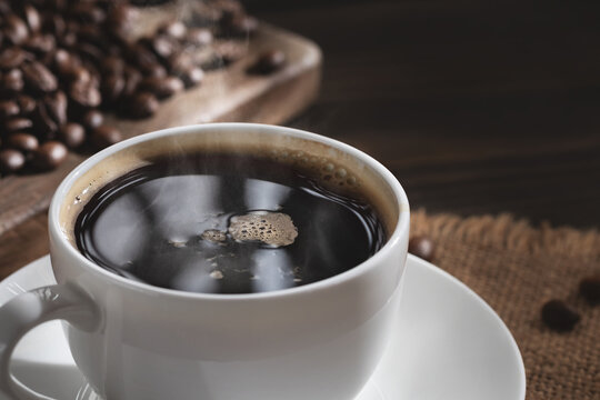 White cup of fresh hot coffee and coffee beans on wooden board on table, close up