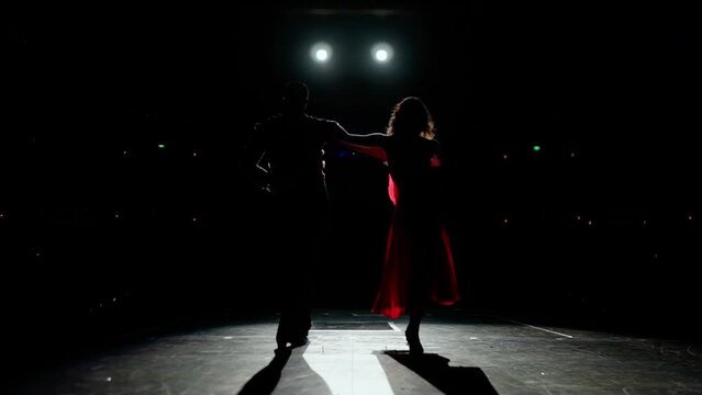 Rear view,man and woman silhouettes starting performance.Ballroom dancers coming out on stage. Scarlet tango dress train develops artistically as butterfly wings