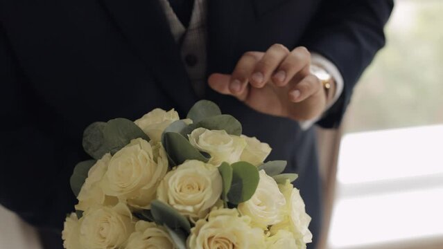 Hands of groom with wedding bouquet at home. White shirt and blue jacket, wrist watch clock. Wedding day. Handsome man preparing to go to bride. Businessman guy. Close-up indoors footage, slow motion