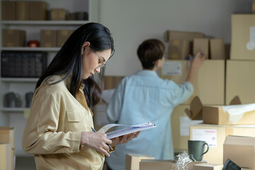 Asian woman online seller confirming orders from customer on the papers. E-commerce business owner working in store warehouse.