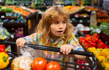 Healthy food for young family with kids. Portrait of smiling little child with shopping cart full of fresh vegetables. Kids at grocery store or supermarket. Shopping cart, grocery store concept.