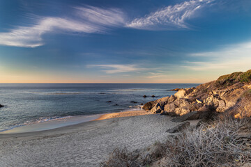 Sandy beach above Malibu, California in early morning. Calm Pacific Ocean in the distance; blue sky and clouds overhead.
