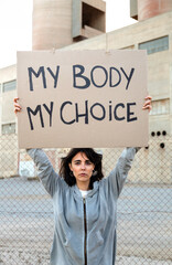 Vertical portrait of young caucasian woman looking at camera holding cardboard sign: My body my...