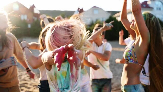 Cheerful girl celebrates Holi festival with friends stained in colored powder. People have fun at hindu holiday. Guys dance, jump on beach in slow motion. End of covid pandemic isolation