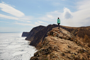 Lake Baikal in spring. White ice floes melt on the water. A woman stands on a rock and enjoys a beautiful view. North of Olkhon Island, Cape Khoboy.