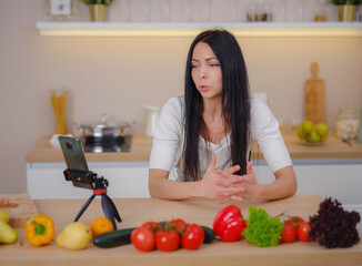 Obraz na płótnie Canvas Professional beautiful happy young woman is blogging for her kitchen channel about healthy living in kitchen of her home and looking on smartphone on tripod