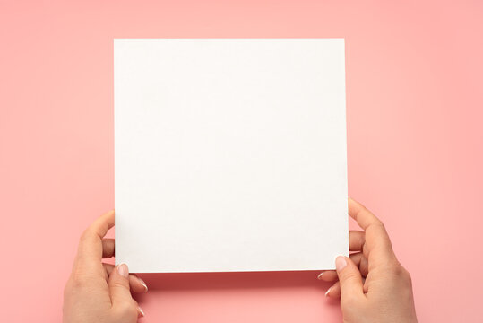 Mockup square paper. Overhead shot of woman hands with blank paper sheet on pink table. Human hands holding mockup white advertising card. Isolated on pink background