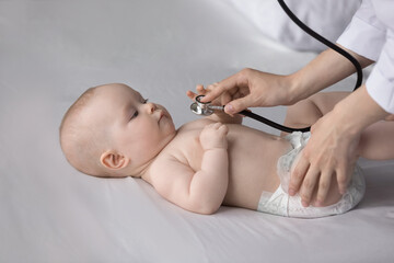 Cropped professional female medical worker pediatrician nurse using stethoscope, listening lungs or heartbeat of happy small newborn baby boy girl in diaper lying on linen at regular checkup meeting.