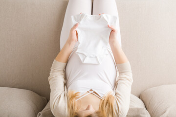 Young pregnant woman with big belly sitting on sofa at living room. Hands holding white bodysuit...