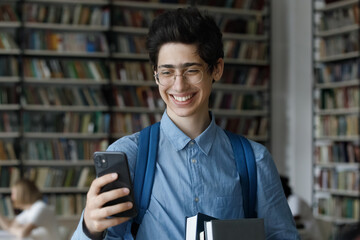 Teenage 17s guy in glasses holds workbooks and smartphone standing in university library, smiling read good news, check message use mobile application. Education, gen Z using modern tech, fun concept