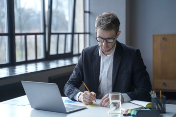 Focused male banker writes down memos on sticky notes while working remotely from home office
