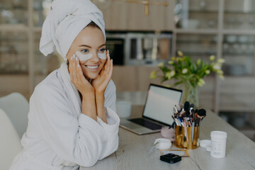 Pretty smiling female model applies collagen patches under eyes to reduce wrinkles wears bathrobe and towel on head enjoys skin care procedures