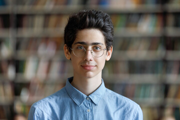 Head shot schoolboy guy posing in campus library on bookshelves background. 17s pupil in eyeglasses...