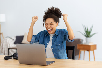 Portrait of excited young African American woman celebrating success while sitting with laptop at...