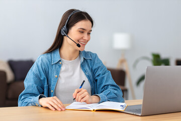 Young smiling freelancer in headset using laptop computer working online from home or modern office. Successful caucasian young female student studying, distance learning, education - obrazy, fototapety, plakaty