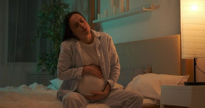 Pregnant woman caressing her belly while suffering with pain sitting on bed at night